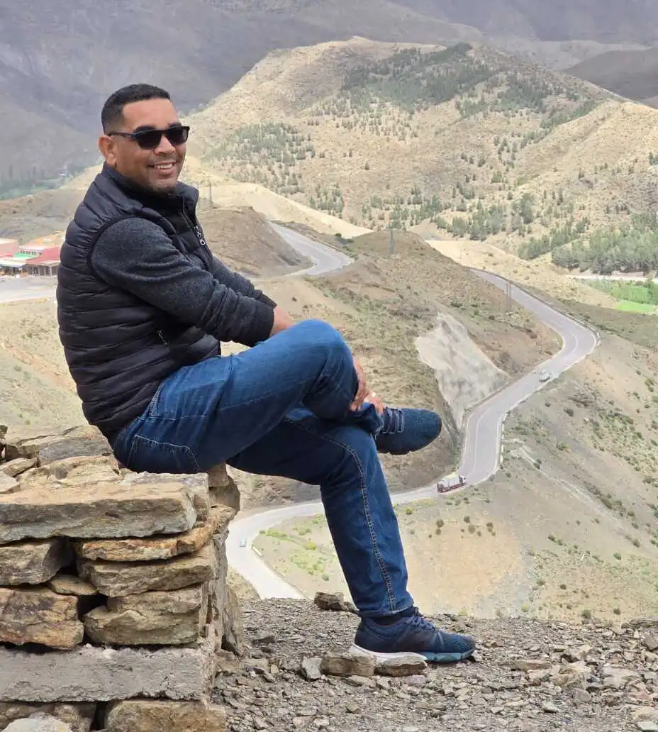 Authentic Morocco Adventures guide sitting and smiling on a mountain viewpoint overlooking winding roads and scenic Atlas Mountains landscape.
